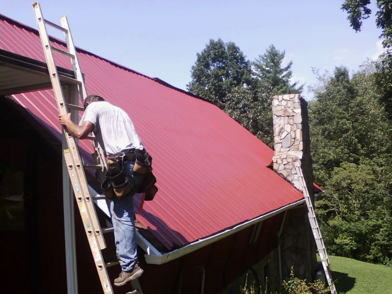 Roofer on a ladder beside a red metal roof and stone chimney — Soffit & Fascia Repair by Roofing Brothers in Rayle